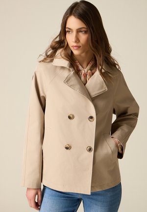 Vrouw draagt een beige dubbelgeknoopte jas over een bloemenblouse, met handen in de zakken, kijkt opzij tegen een effen achtergrond.