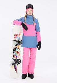 Snowboarder con un traje azul y rosa, sosteniendo una tabla de snowboard con gráficos coloridos. Incluye un cuello alto, manoplas y gafas de esquí.