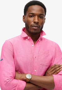 Camisa de botón de color rosa claro con un bolsillo en el pecho, mangas enrolladas y una textura sutil. Presenta un pequeño parche con el logo en el brazo izquierdo.