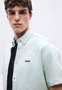 Νέος άνδρας που φοράει πουκάμισο με κοντό μανίκι και κουμπιά σε ανοιχτό πράσινο χρώμα με την ένδειξη "BOSS" κεντημένη στο στήθος και μαύρη μπλούζα από μέσα.