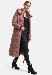 Long pink puffer coat with a zip closure and hood, featuring a quilted pattern and side zip pockets. Worn over black jeans and heeled boots.