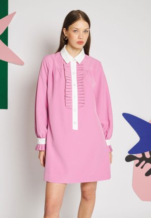 Vrouw draagt een roze jurk met lange mouwen, met een witte kraag en manchetten, staand tegen een witte achtergrond met abstracte vormen.