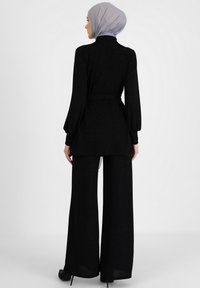 Completo nero con una tunica a maniche lunghe, con cintura, in tessuto testurizzato e pantaloni larghi. Gli accessori includono un hijab grigio chiaro.