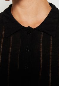 Czarna odzież z kołnierzykiem, z pionowymi paskami. Posiada małe czarne guziki oraz teksturowany materiał o lekko prześwitującym wyglądzie.