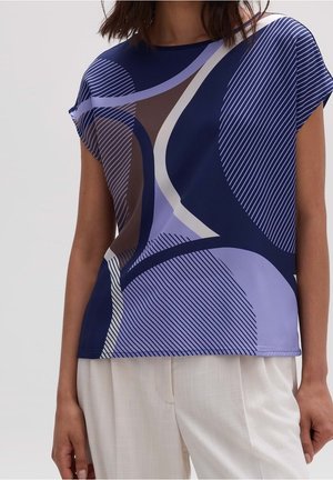 Woman wearing a short-sleeve top with abstract blue, lavender, brown, and white curved and striped patterns, paired with cream-colored pants.