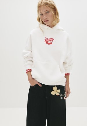 Jeune femme portant un sweat à capuche blanc surdimensionné avec un graphique de cerise rouge et un pantalon large noir avec un nœud et un accessoire en chaîne.
