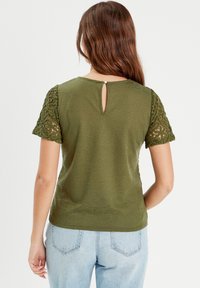 Haut vert olive avec des manches courtes en dentelle et un détail en trou de serrure au dos. Le tissu est texturé et légèrement ample.