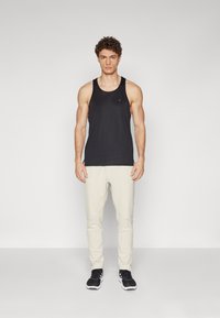 Black sleeveless tank top, fitted design, with a round neck and subtle logo; paired with light beige tapered pants and black sneakers.