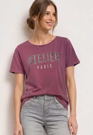 Kobieta z jasnobrązowymi włosami w niskim koku ma na sobie fioletowy t-shirt z napisem "Atelier de Paris" i jasnoszare dżinsy, uśmiecha się i patrzy w prawo.