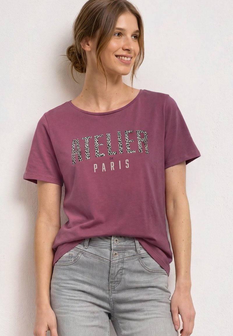 Femme aux cheveux châtain clair en chignon bas porte un t-shirt violet avec le texte "Atelier de Paris" et un jean gris clair, souriante et regardant vers la droite.