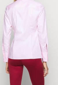 Donna che indossa una camicia a maniche lunghe aderente di un rosa chiaro e pantaloni aderenti borgogna, vista da dietro su uno sfondo semplice.