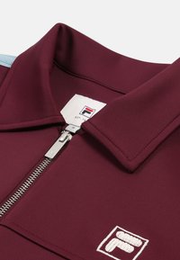 Polo burdeos con cremallera y un acento en azul claro. Tiene un cuello plano y una cremallera plateada. Parche del logotipo cosido en el área del pecho.