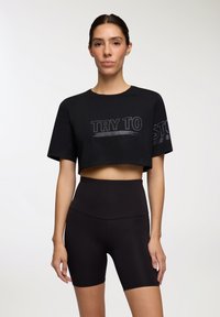 Czarna, krótka koszulka z napisem "SPRÓBUJ ZATRZYMAĆ" w błyszczącym wykończeniu, zestawiona z czarnymi, wysokimi szortami rowerowymi. Gładka faktura, casualowy design.