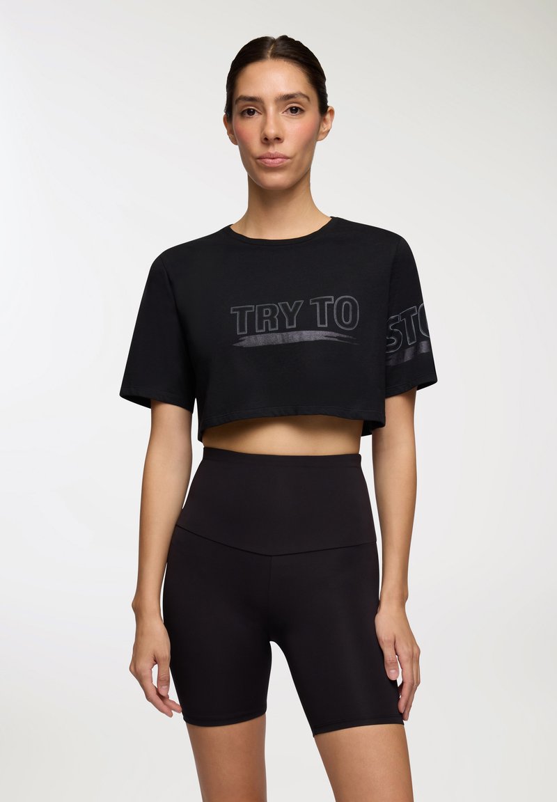 Czarna, krótka koszulka z napisem "SPRÓBUJ ZATRZYMAĆ" w błyszczącym wykończeniu, zestawiona z czarnymi, wysokimi szortami rowerowymi. Gładka faktura, casualowy design.