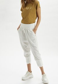 T-shirt en coton marron avec "Unique" imprimé en or, associé à un pantalon de jogging blanc avec une taille élastique et des poignets, porté avec des baskets blanches.