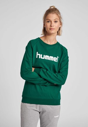 Lyshåret kvinde med håret i en topknude iført en grøn sweatshirt og grå joggingbukser, begge med "hummel"-logoer, står med armene krydset.