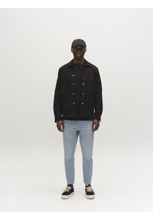 Férfi áll, fekete sapkát, fekete gombos dzsekit, fekete inget, világoskék farmert és fekete tornacipőt visel fehér talppal és fűzővel.