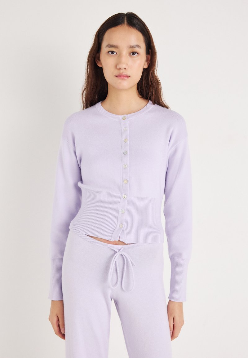 Κοντό λιλά ζακέτα με μακριά μανίκια, ριμπ τελείωμα και κουμπιά στο μπροστινό μέρος. Η απαλότητα της υφής ταιριάζει με παντελόνια με ρίγα και κορδόνι στη μέση.