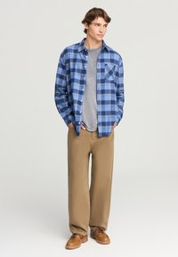 Giovane uomo che indossa una camicia a quadri blu sopra una maglietta grigia, pantaloni beige e scarpe marroni, in piedi con le mani nelle tasche, che guarda a destra.