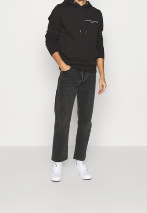 Persona che indossa un hoodie nero di Tommy Hilfiger, jeans grigio scuro e sneakers bianche, in piedi con una mano in tasca.