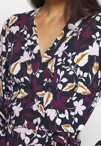 Květinové šaty s hlubokým V-výstřihem, vyrobené z tmavé tkaniny se vzorem fialových, růžových a bílých květin, a zlatými akcenty ve tvaru listů.