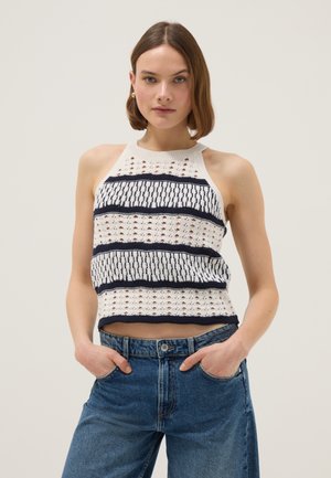 Mujer con cabello castaño corto, vistiendo una blusa sin mangas de crochet blanca con rayas azules marinas y jeans de mezclilla azul, con las manos en los bolsillos, fondo neutro.