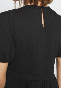 Vue de dos d'une personne portant une robe noire avec des manches courtes bouffantes et une petite ouverture en forme de goutte au niveau du haut du dos.