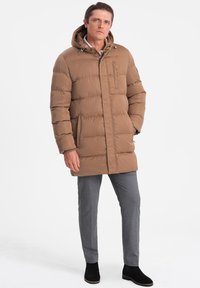 Giacca a vento marrone con cappuccio, chiusura zip e tasca sul petto. Presenta pannelli cuciti e tasche laterali. Abbinata a pantaloni grigi e stivali neri.