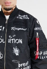 Black bomber jacket with white text graphics, zip closure, ribbed collar, and a red tag on the sleeve reading "REMOVE BEFORE FLIGHT."