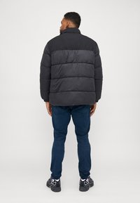 Sort polstret jakke med høj krave og vandret quilting, parret med blå jeans og sorte sneakers, set fra ryggen.