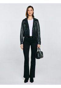 Sieviete tērpusies melnā ādas jaku, baltā kreklā, melnās platajās biksēs un melnos zābakos, turot melnu rokassomiņu uz vienkārša fona.