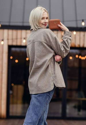 Beige, oversized, wool-blend shirt with a curved hem, worn over blue jeans. The model holds a small brown wallet, displaying a logo.