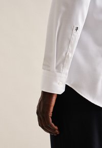 Camicia bianca a maniche lunghe con una texture liscia, caratterizzata da un polsino con bottoni e una piccola rosa nera ricamata sulla manica.