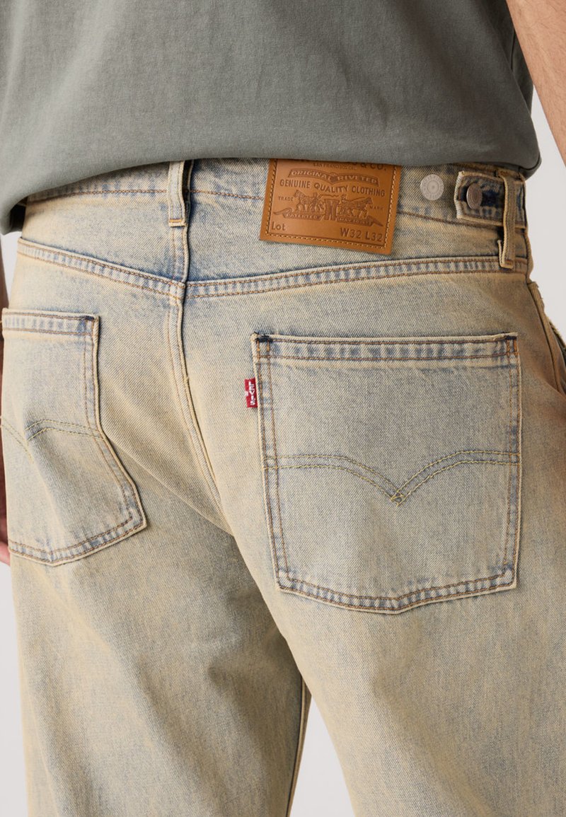 Jeans in denim azzurro chiaro con finitura sbiadita, dotati di due tasche posteriori, cuciture a contrasto e una toppa in pelle marrone sulla cintura.