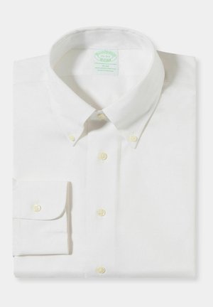 Bílá úzká košile s dlouhým rukávem, límečkem na knoflíky a knoflíky na manžetách, pečlivě složená na jednobarevném pozadí.