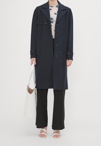Trench coat azul marino de corte holgado, con gran solapa y botones al frente, combinado con pantalones negros de pierna ancha y sandalias blancas con tiras, sosteniendo un bolso blanco.
