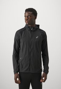 Atleetlik mees, kes kannab musta kapuutsiga lukuga jakki ja musti pükse, vaatab vasakule tavalise hele tausta taustal.