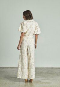 Camicia color crema e pantaloni larghi con motivi intricati. Presenta maniche a sbuffo e una vita arricciata, il materiale sembra testurizzato.