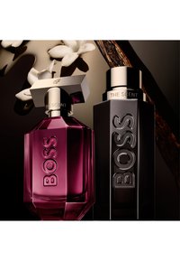 Twee Boss The Scent parfumflesjes, één roze en gefacetteerd, één zwarte en rechthoekig, met witte bloemen en vanilleboon op de achtergrond.