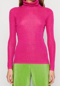 Žena oblečená v lesklém růžovém roláku v kombinaci s jasně zelenými sklady kalhot, stojící na jednobarevném pozadí.