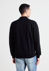 Svart sweatshirt med høy hals, laget av mykt stoff. Har ribbet kant og mansjetter, kombinert med lyseblå denimjeans.