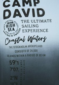 Tela de color azul claro con texto impreso en negro que destaca "Camp David," experiencias de navegación y coordenadas geográficas, con acentos decorativos dispersos.