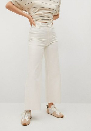 Personne portant un jean blanc taille haute à jambes larges, un top à manches courtes rayé, et des baskets beige et blanches, debout les mains sur les hanches.