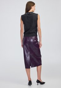 Kobieta stoi, ubrana w bezrękawnik w ciemnoszarym kolorze z ozdobnym kołnierzykiem oraz błyszcząca, ciemnofioletowa skórzana spódnica midi z rozporkiem z tyłu.