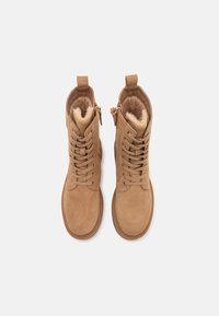 Tan värvi nubukjalatsid pahkluuni, millel on nööridega esiosa, külgkaTask ja pehme karvane vooder. Ümar varvas ja õmmeldud detailid talla äärtes.