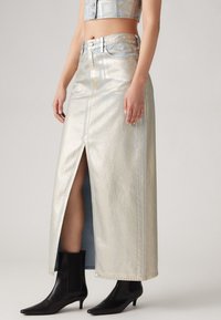 Fustă din denim metalizat de culoare albastru deschis, cu o fanta înaltă și cusături contrastante, asortată cu botine negre cu vârful ascuțit.