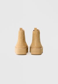 Une paire de bottes à plateforme en suède beige vue de dos avec des languettes de tirage et des détails cousus sur un fond gris uni.