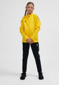 Gul zip-up jacka med ståkrage, kombinerad med svarta träningbyxor med chevronmönster längs sidorna. Svarta sneakers fulländar outfiten.