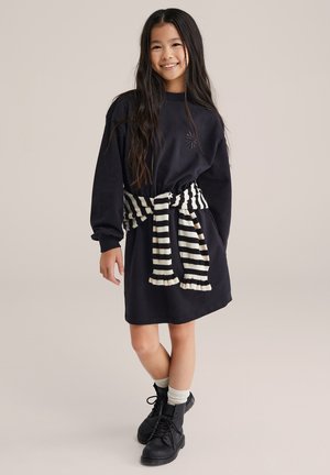 Tengerészkék pulóverruha hímzett mintával, fekete-fehér csíkos, övként viselt sállal a deréknál megkötve, fekete csizmával párosítva.