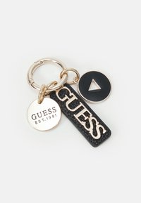 Nøglebundt med guldring og tre vedhæng: rundt guldmærke med teksten "GUESS EST. 1981," sort rektangel med guld "GUESS," og sort cirkel med guldtriangle.